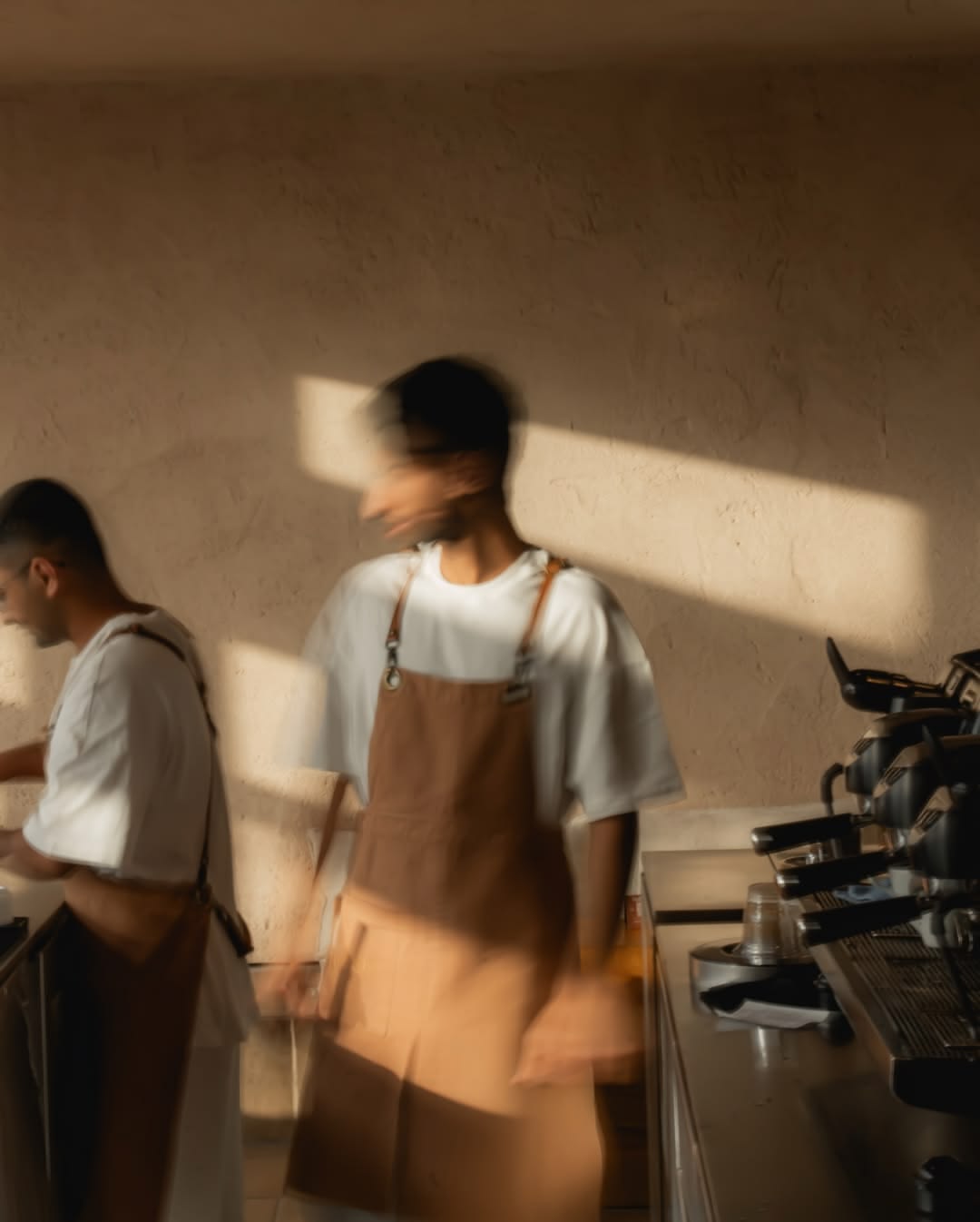 Two baristas in white tees and tan canvas aprons at the espresso bar, softly blurred with motion, warm late-afternoon sun striping the plaster wall behind them.