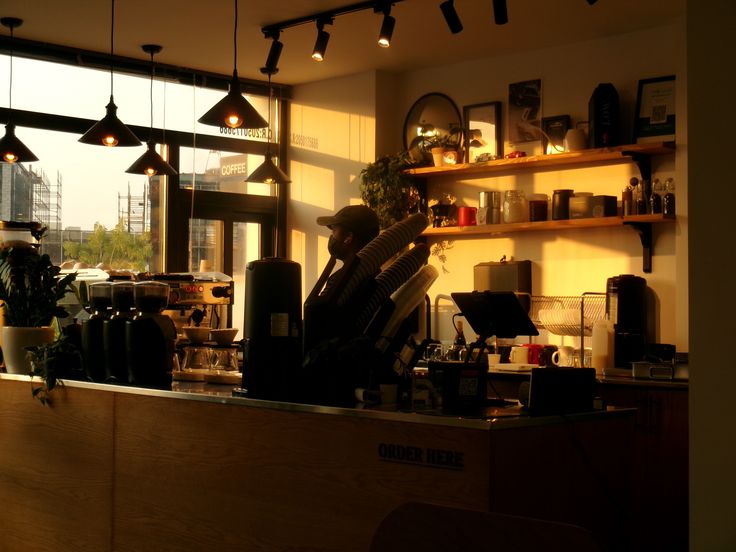 Golden-hour light pouring through the front windows onto the coffee bar: a barista in a cap works behind the espresso machine, pendant lamps hang overhead, and a wooden shelf of cups and jars runs along the back wall.