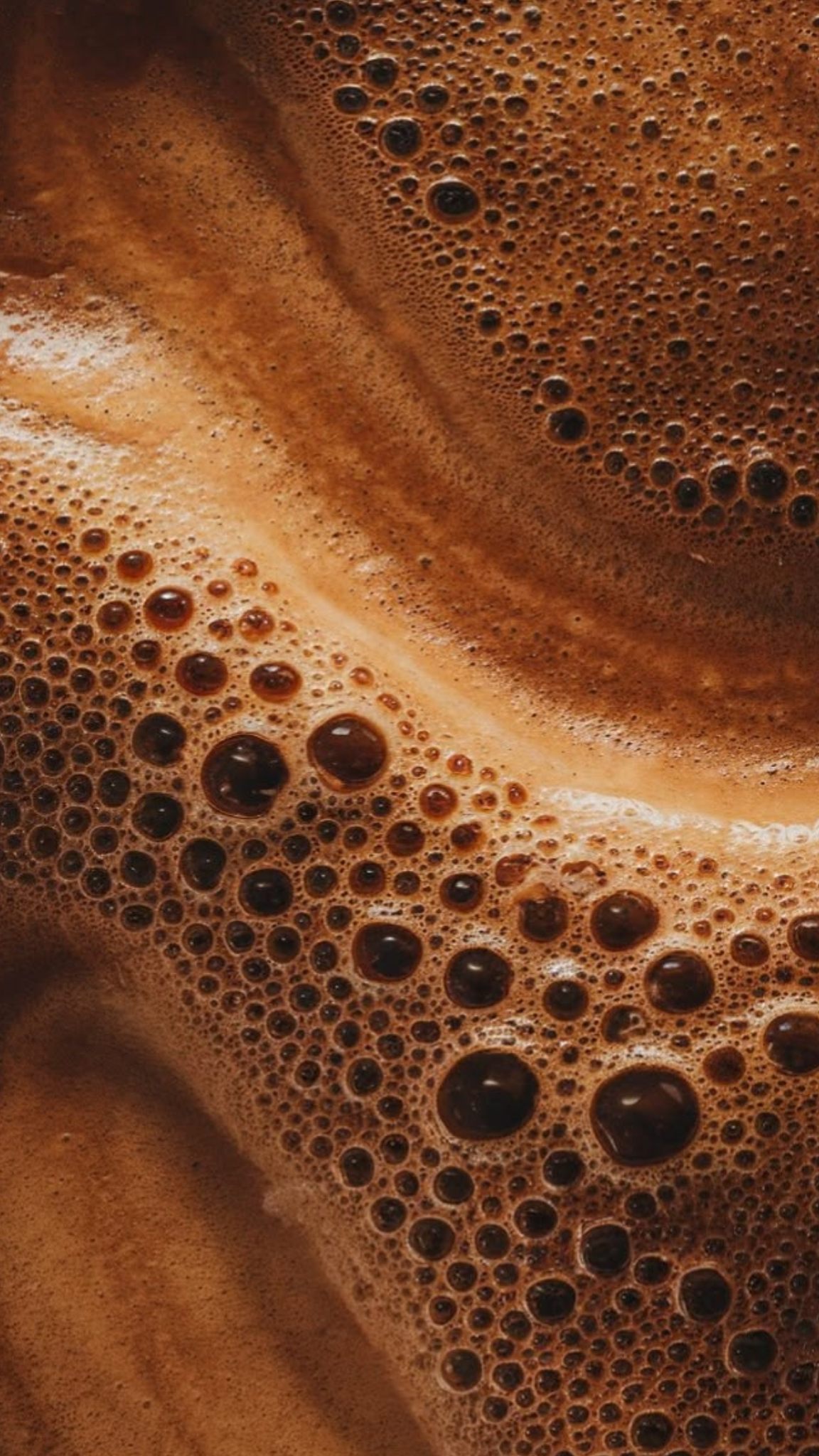 Abstract close-up of golden-brown coffee crema, its surface scattered with tiny bubbles and slow swirls.