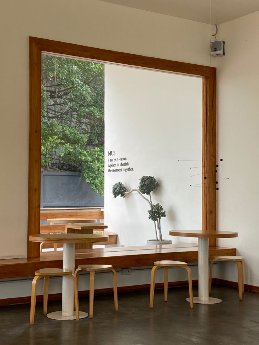 A tall teak-framed window looking onto green trees, with a low wooden bench, two round tables and pale stools set in front of it.