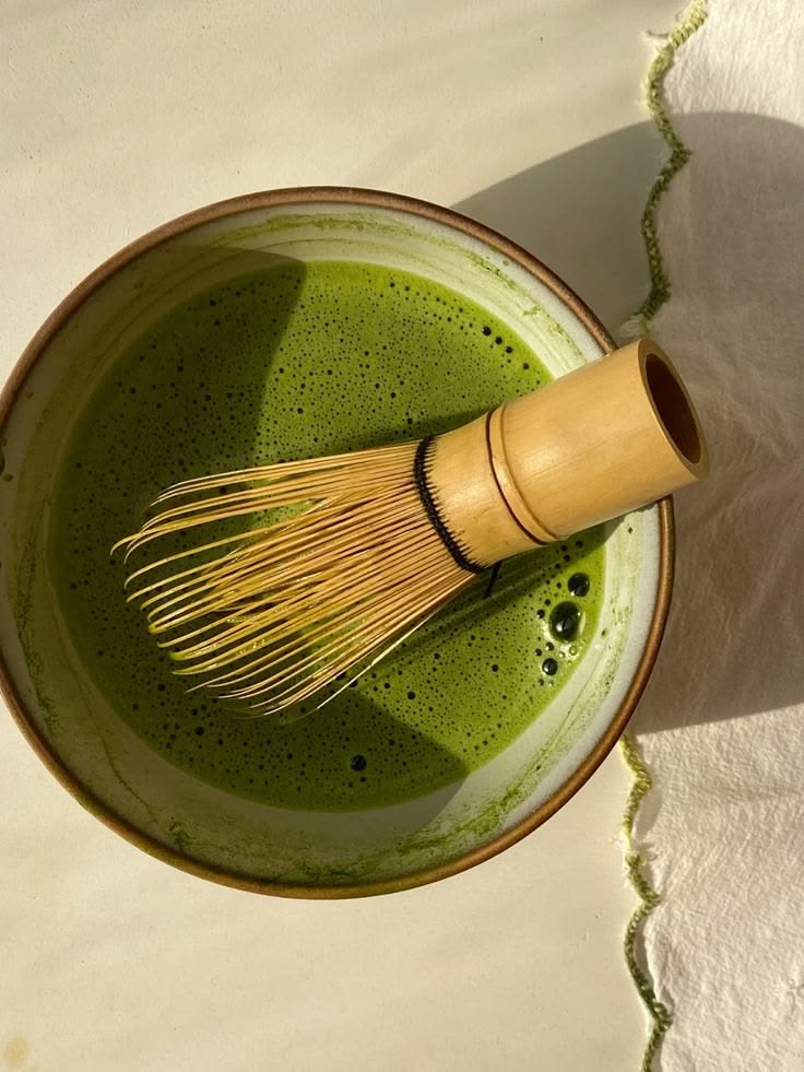 A bamboo whisk resting in a ceramic bowl of freshly whisked matcha, bright green with fine foam, on a white linen cloth.