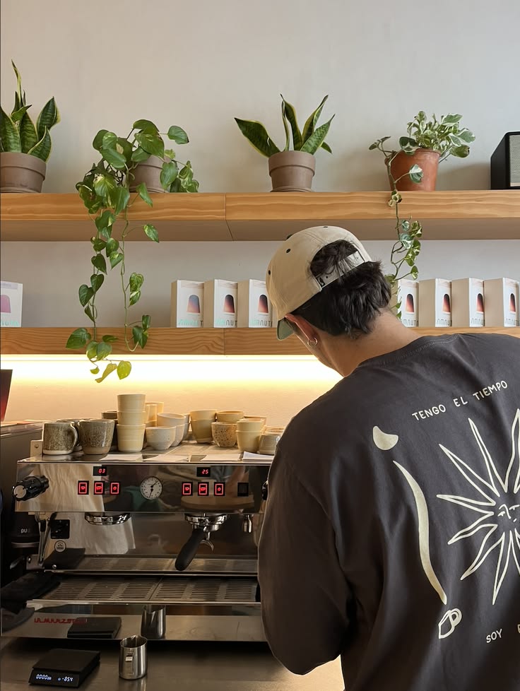 A barista in a cap, seen from behind, working at the espresso machine beneath a wooden shelf of trailing plants and stacked cups.