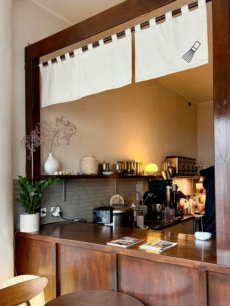 A dark wooden counter beneath a white noren curtain, with a small vase of dried flowers, a paper lantern and a shelf of ceramics behind the bar.