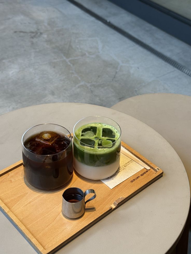 An iced black coffee and an iced matcha latte in short glasses on a wooden tray, with a small steel jug of espresso beside them.