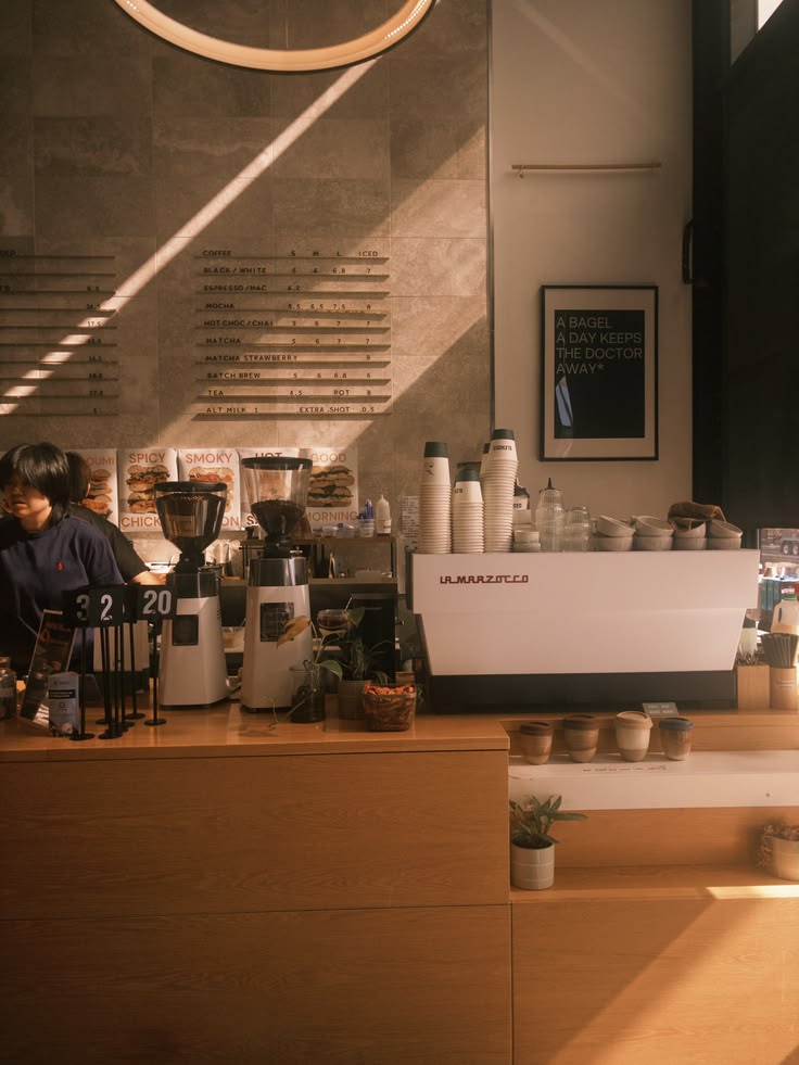 Late sun striping a stone wall above the bar, where a barista works beside two grinders, a white espresso machine and rows of stacked cups.