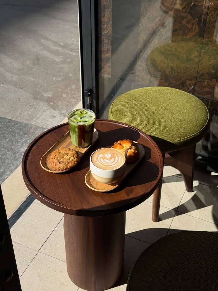 A small round walnut table in the window with a flat white, an iced matcha, a cookie and a muffin on wooden trays, beside a green upholstered stool.