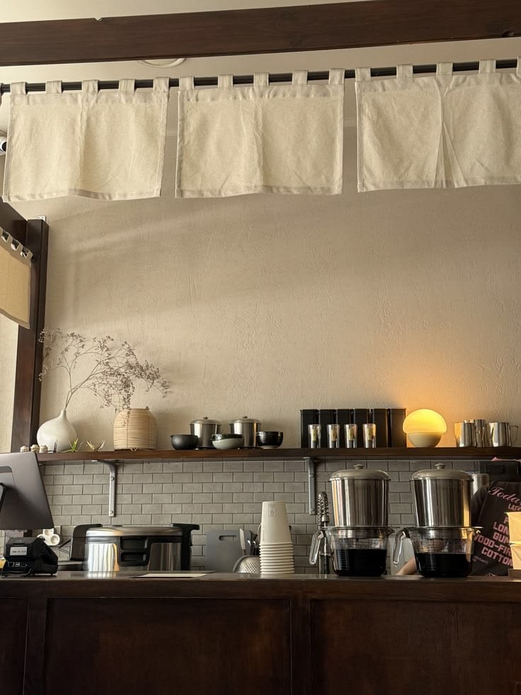 White linen curtains hung along a beam above a plaster wall, a shelf of ceramics and a glowing lamp, and two steel brewers on the counter below.