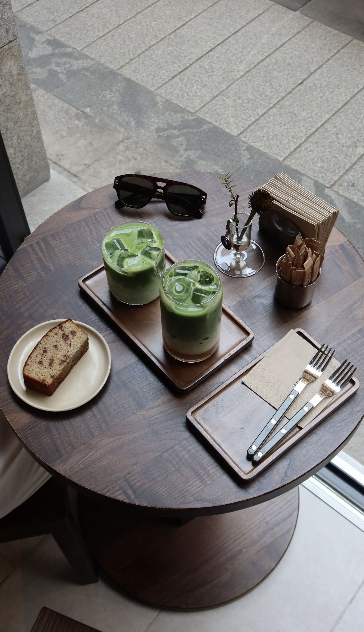 Seen from above: two iced matcha lattes on a wooden tray, a slice of banana bread on a cream plate, sunglasses and cutlery on a round dark-wood table.