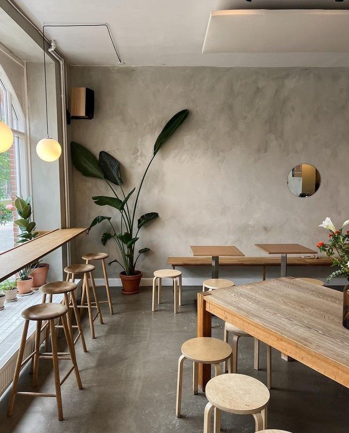 The seating area at Fuku Coffee: light oak stools along a window counter, a long wooden communal table, plaster walls, a round mirror and a tall bird-of-paradise plant in soft daylight.